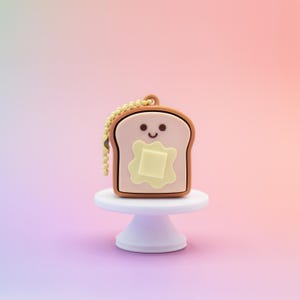 May include: A cute, cartoon-style keychain shaped like a slice of toast with a pat of butter. The toast has a smiling face and is on a small white pedestal. The keychain has a gold-colored chain and a brown border.