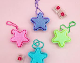 May include: An assortment of star-shaped keychains in pink, blue, green, and turquoise, each with a matching beaded chain. Two small, clear square objects with a red plus sign are also present. The background is a solid pink.