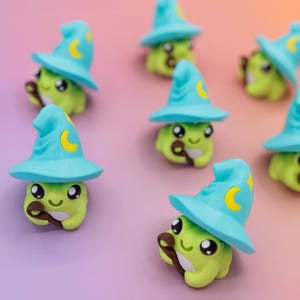 May include: A collection of whimsical green frog figurines, each wearing a light blue wizard hat with a yellow crescent moon. The frogs hold a brown wand and have large, expressive eyes. The background is a gradient of purple and orange.