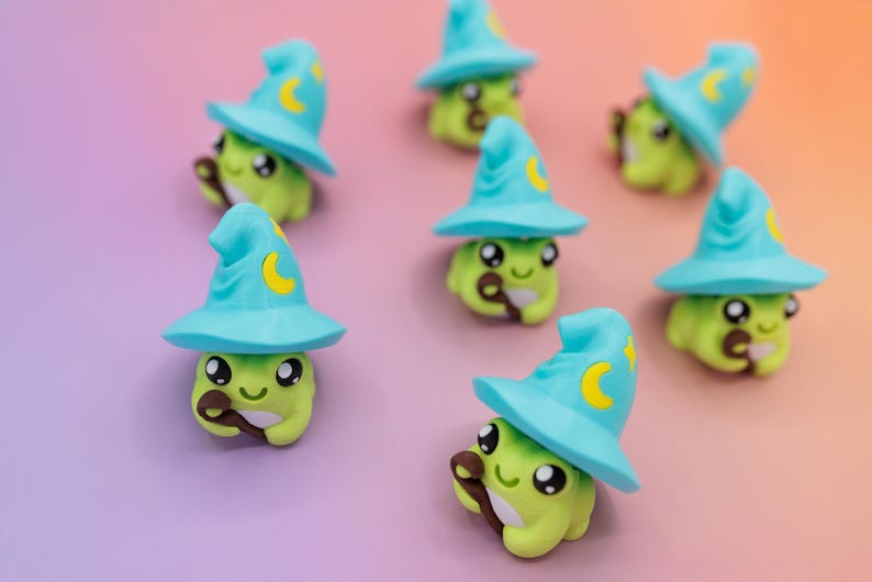 May include: A collection of whimsical green frog figurines, each wearing a light blue wizard hat with a yellow crescent moon. The frogs hold a brown wand and have large, expressive eyes. The background is a gradient of purple and orange.
