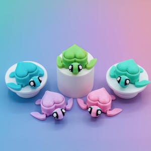 May include: A collection of colorful turtle-shaped figurines. The turtles are in shades of blue, green, and pink, with white accents. Some are on white platforms, while others are free-standing. The background is a gradient of blue and pink.