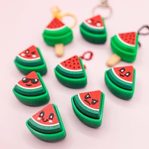 May include: A collection of watermelon-themed keychains and charms. The charms are shaped like watermelon slices and popsicles, featuring red, green, and white colors. Some have smiling or angry faces. The background is a soft pink.