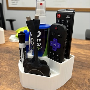 May include: A white desk organizer with five compartments holds a variety of items, including a black remote control, a purple remote control, a black and white marker, a black and white cable, a black and white device, and a blue spray bottle with a white cap. The bottle has the text "White Board Cape" on it.