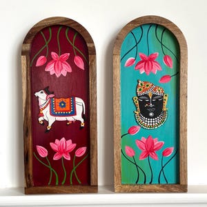 Peut inclure: Deux œuvres d'art murales en bois. L'une représente une vache blanche avec une couverture colorée sur un fond bordeaux avec des fleurs de lotus roses. L'autre représente une divinité noire avec une couronne ornée de bijoux sur un fond turquoise avec des fleurs de lotus roses.