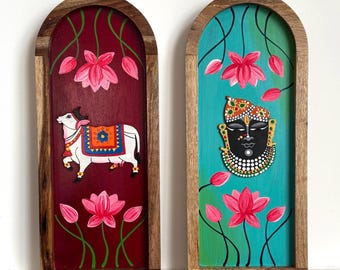 May include: Two arched wooden wall plaques featuring hand-painted designs. The left plaque has a deep red background with a white cow and pink lotus flowers. The right plaque has a teal background with a black deity face and pink lotus flowers.