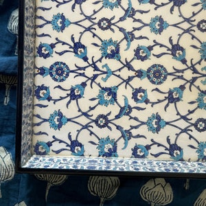May include: A rectangular serving tray with a black frame. The tray's surface features a repeating floral pattern in shades of blue and turquoise on a white background. The floral design is reminiscent of traditional tile work.