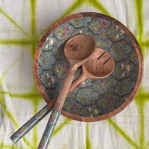 Op de afbeelding: Een houten slakom met een turquoise bloemenpatroon en twee houten saladelepels. De kom heeft een natuurlijke houten rand en de lepels hebben turquoise handvatten met een bloemenpatroon. De achtergrond is een witte doek met een groen tie-dye patroon.