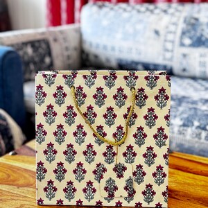 May include: A square gift bag with a cream-colored background and a repeating pattern of maroon floral designs. The bag has a gold-colored rope handle and a matching tag. The bag is sitting on a wooden surface.