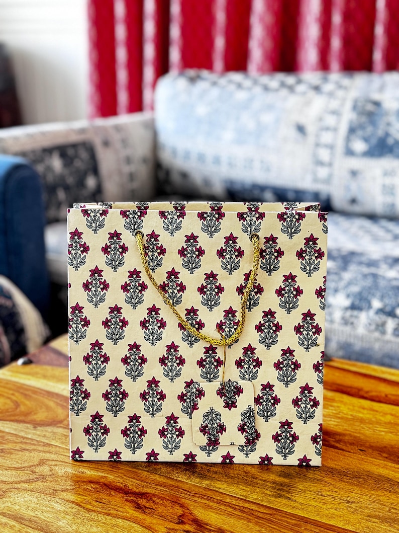 May include: A square gift bag with a cream-colored background and a repeating pattern of maroon floral designs. The bag has a gold-colored rope handle and a matching tag. The bag is sitting on a wooden surface.