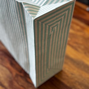 May include: A rectangular gift bag with a vertical striped pattern in light blue and white. The bag is made of paper and has a folded top and bottom. The bag is sitting on a wooden surface.