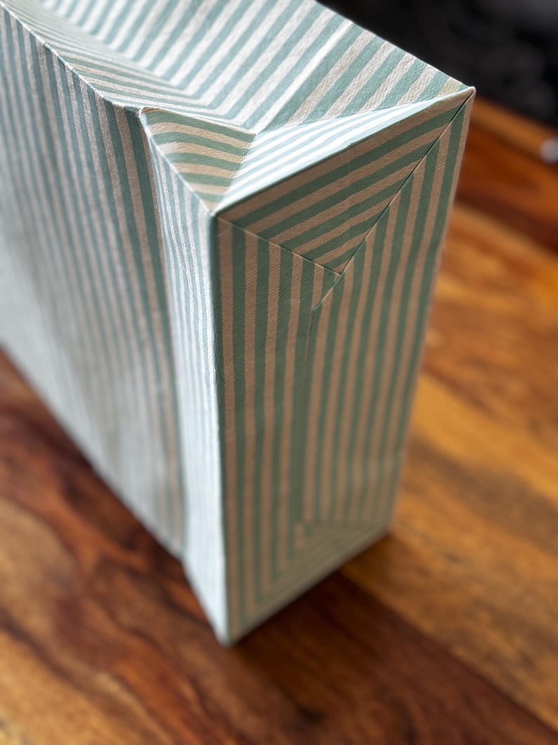 May include: A rectangular gift bag with a vertical striped pattern in light blue and white. The bag is made of paper and has a folded top and bottom. The bag is sitting on a wooden surface.