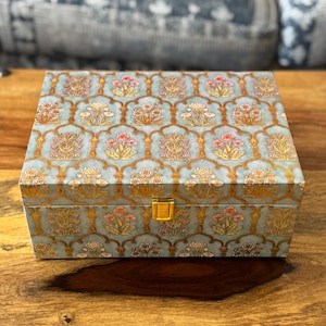 Puede incluir: Una caja decorativa cuadrada con un estampado floral en tonos azules, rosas y dorados. La caja tiene un cierre y adornos dorados. La caja está sobre una superficie de madera.