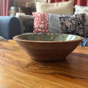 Op de afbeelding: Een houten kom met een lichtbruine buitenkant en een bloemmotief aan de binnenkant. De kom staat op een houten tafel. De binnenkant van de kom heeft een lichtblauwe achtergrond met een bloemmotief in roze, groene en gele tinten.