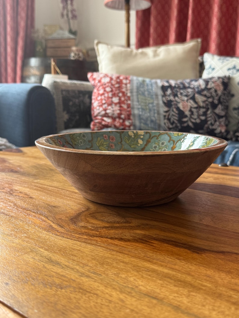 Op de afbeelding: Een houten kom met een lichtbruine buitenkant en een bloemmotief aan de binnenkant. De kom staat op een houten tafel. De binnenkant van de kom heeft een lichtblauwe achtergrond met een bloemmotief in roze, groene en gele tinten.