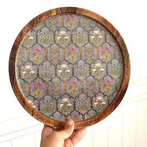 May include: A round wooden serving tray featuring a light blue interior surface with a repeating pattern of arched frames containing small floral bouquets in shades of pink, white, and green. The tray has a smooth, dark brown natural wood rim.