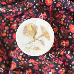 May include: A round, white marble trinket box lid with a floral design. The flower and leaves are made of inlaid, iridescent, golden-colored shell pieces. The lid is set against a dark background with a floral pattern in shades of red, pink, and orange.