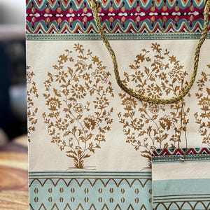 May include: Cream-colored gift bag with a gold rope handle. The bag features a repeating pattern of gold floral trees and decorative borders in teal, red, and gold. The bag is made of paper and is suitable for gifts.