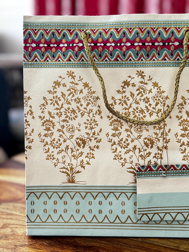May include: Cream-colored gift bag with a gold rope handle. The bag features a repeating pattern of gold floral trees and decorative borders in teal, red, and gold. The bag is made of paper and is suitable for gifts.