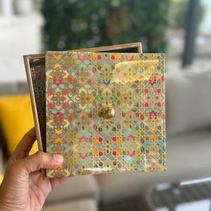 Op de afbeelding: Een vierkante houten doos met een deksel voorzien van een kleurrijk geometrisch patroon in mintgroen, geel, roze en rood. Het deksel heeft een kleine, centraal geplaatste goudkleurige knop. De doos staat open en toont een donkere binnenzijde.