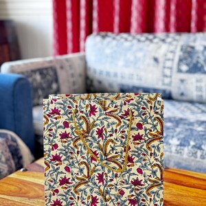 May include: A square paper gift bag with a floral pattern in cream, burgundy, and gold. The bag features a repeating design of stylized flowers and leaves. It has gold rope handles and is sitting on a wooden table.