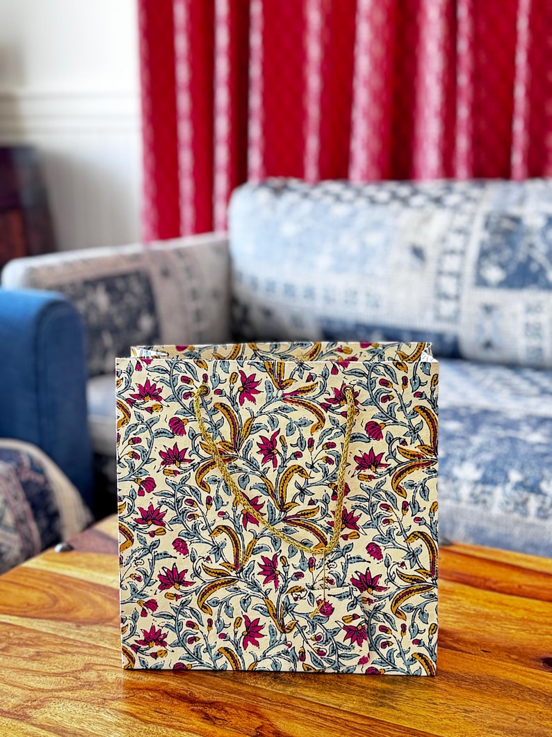 May include: A square paper gift bag with a floral pattern in cream, burgundy, and gold. The bag features a repeating design of stylized flowers and leaves. It has gold rope handles and is sitting on a wooden table.
