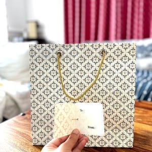 May include: A patterned gift bag with a gold rope handle. The bag has a cream background with a repeating pattern of gray and gold floral shapes. A small white gift tag is held in front of the bag, with the words "To:" and "From:" printed on it.