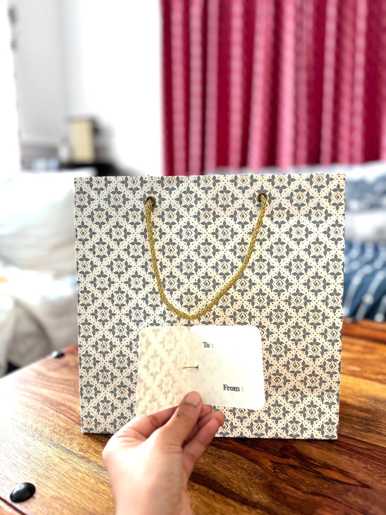 May include: A patterned gift bag with a gold rope handle. The bag has a cream background with a repeating pattern of gray and gold floral shapes. A small white gift tag is held in front of the bag, with the words "To:" and "From:" printed on it.