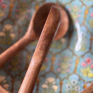 Op de afbeelding: Close-up van een houten saladelepel met een afgerond uiteinde, tegen een decoratieve kom. De kom heeft een lichtblauwe achtergrond met een bloemenpatroon in roze, geel en wit.