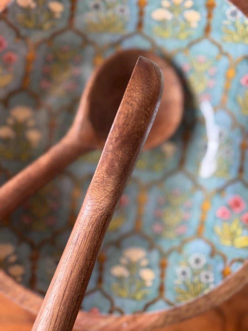 Op de afbeelding: Close-up van een houten saladelepel met een afgerond uiteinde, tegen een decoratieve kom. De kom heeft een lichtblauwe achtergrond met een bloemenpatroon in roze, geel en wit.