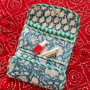 Può includere: Un beauty case trapuntato con stampa floreale verde acqua e bianca, con chiusura a scatto. La borsa è aperta, rivelando uno smalto rosso e un rossetto. La borsa è rifinita con tessuto a righe verdi e bianche.