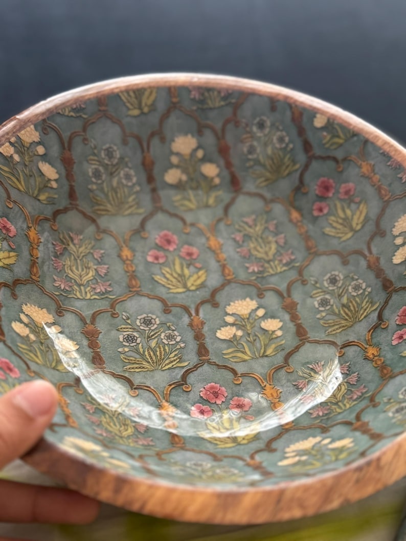 Op de afbeelding: Een decoratieve kom met een turquoise interieur met een bloemenpatroon in roze, geel en wit. De rand is van natuurlijk hout. Het ontwerp van de kom bevat een ingewikkeld, zich herhalend boogmotief. Diameter ongeveer 20 cm.