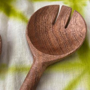 Op de afbeelding: Een houten saladelepel met een brede, afgeronde kop en twee sleuven. De lepel is gemaakt van lichtbruin hout en heeft een lange steel. De lepel is ontworpen voor het serveren van salades en andere gerechten.