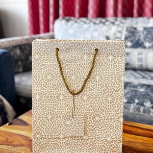 May include: A beige gift bag with a repeating white geometric pattern. The bag has a gold rope handle and a small tag. The bag is sitting on a wooden table.