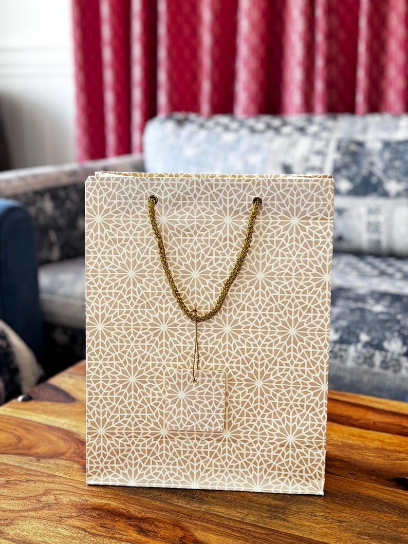 May include: A beige gift bag with a repeating white geometric pattern. The bag has a gold rope handle and a small tag. The bag is sitting on a wooden table.