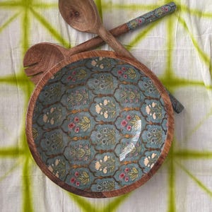 Op de afbeelding: Een houten slakom en serveerlepels. De kom heeft een turquoise bloemenpatroon en een natuurlijke houten rand. De lepels zijn van hout met bloemaccenten op de handvatten. De achtergrond is een wit en groen tie-dye stof.