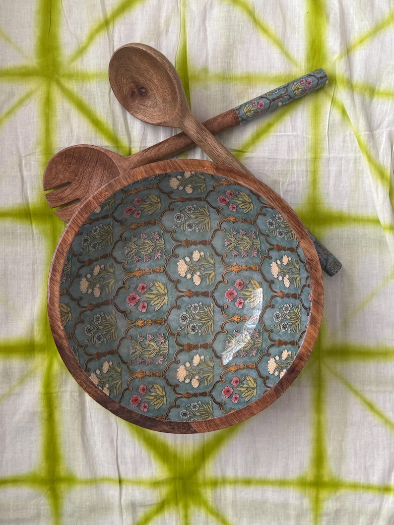 Op de afbeelding: Een houten slakom en serveerlepels. De kom heeft een turquoise bloemenpatroon en een natuurlijke houten rand. De lepels zijn van hout met bloemaccenten op de handvatten. De achtergrond is een wit en groen tie-dye stof.