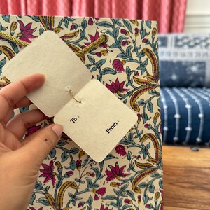 May include: Gift bag with a floral pattern in pink, yellow, and teal on an off-white background. The bag has a gold rope handle and a tag that says "To:" and "From:"