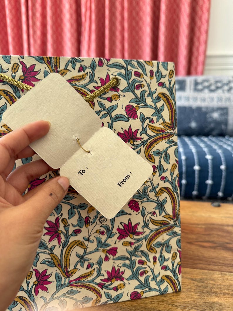 May include: Gift bag with a floral pattern in pink, yellow, and teal on an off-white background. The bag has a gold rope handle and a tag that says "To:" and "From:"