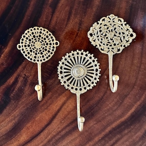 May include: Three decorative brass wall hooks with detailed openwork patterns. The hooks display circular, sunburst, and floral designs. Each hook has a curved lower section for hanging items.