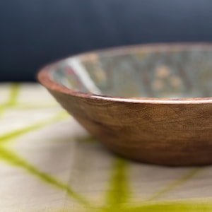 Op de afbeelding: Een houten kom met een natuurlijke afwerking. De kom heeft een brede, ondiepe vorm en een glad, gepolijst oppervlak. De binnenkant van de kom heeft een lichtgroen en bruin patroon. De kom staat op een wit en groen patroon doek.