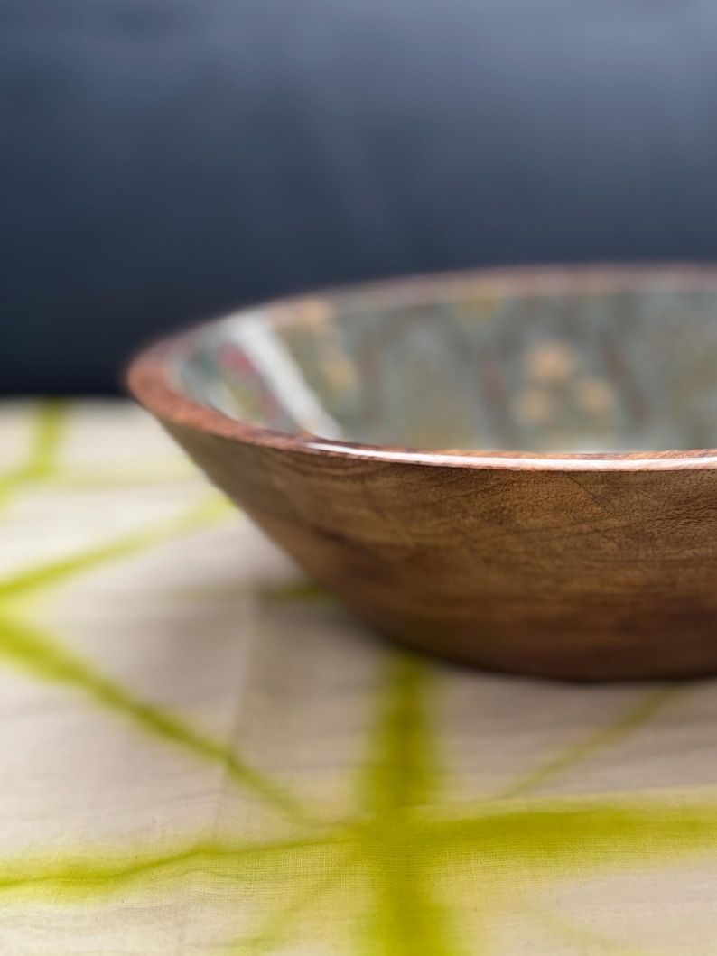 Op de afbeelding: Een houten kom met een natuurlijke afwerking. De kom heeft een brede, ondiepe vorm en een glad, gepolijst oppervlak. De binnenkant van de kom heeft een lichtgroen en bruin patroon. De kom staat op een wit en groen patroon doek.