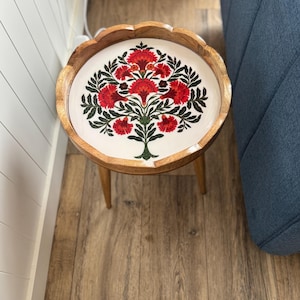 Può includere: Un tavolino rotondo in legno con bordo smerlato e tre gambe affusolate. Il piano del tavolo presenta una superficie bianca decorata con un'illustrazione botanica centrale di un albero con foglie verde scuro e fiori rossi vivaci.
