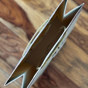 May include: An overhead view of a gift bag with a gold interior and a gold rope handle. The bag is light beige with gold trim and a floral design. The bag is open, revealing the inside.