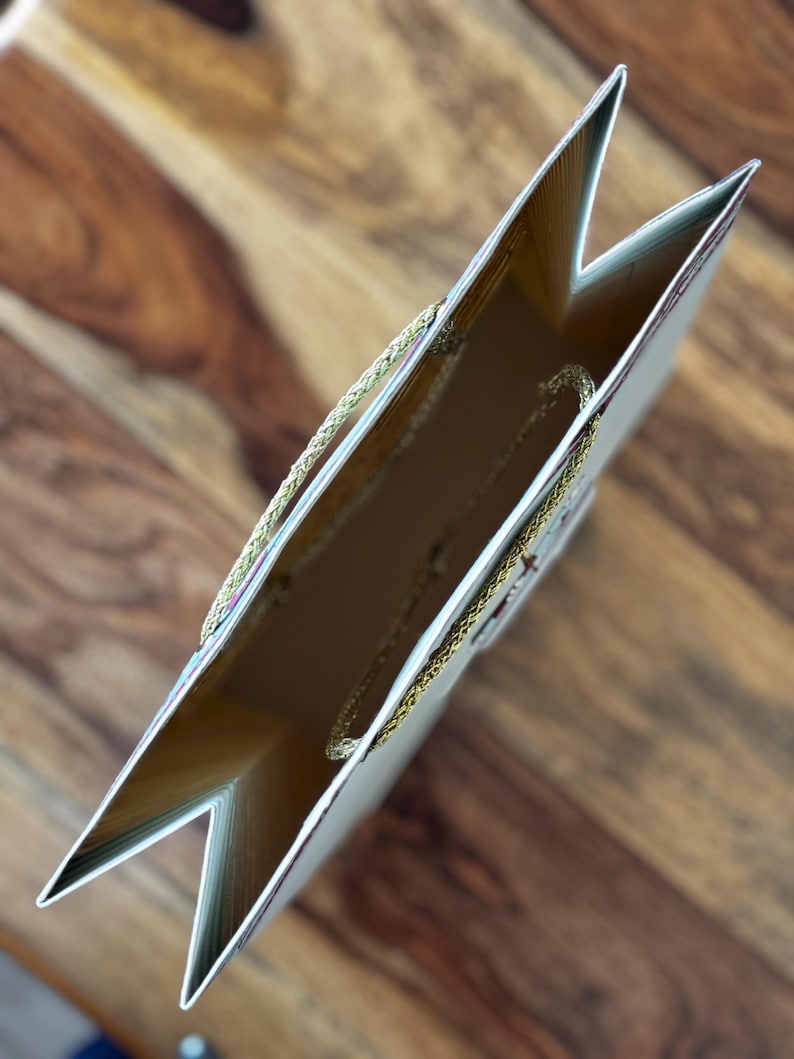May include: An overhead view of a gift bag with a gold interior and a gold rope handle. The bag is light beige with gold trim and a floral design. The bag is open, revealing the inside.