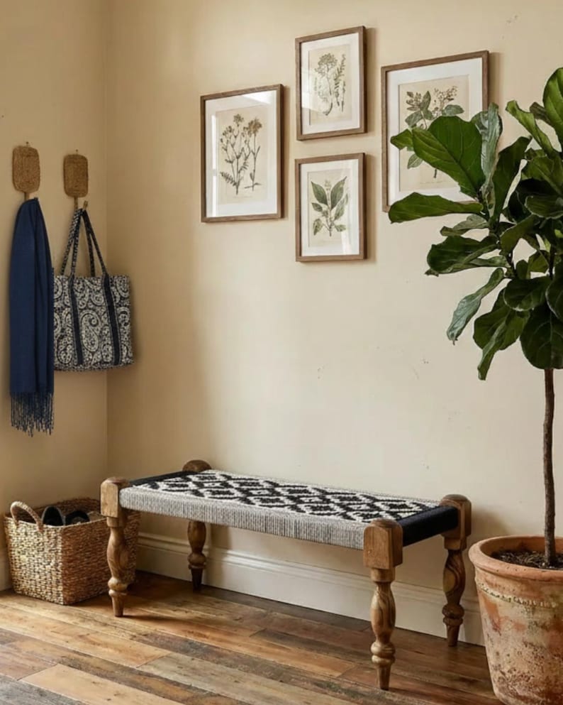 May include: A room corner with a wooden bench with a black and white patterned seat, framed botanical prints, and a potted plant. A blue scarf and patterned tote bag hang on the wall, next to a woven basket.