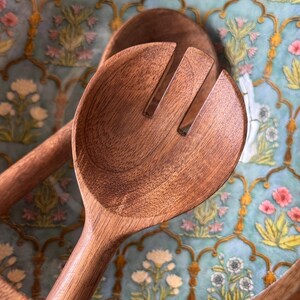 Op de afbeelding: Houten saladebestek met een sleuflepel en vork rust op een decoratieve kom. De kom heeft een lichtblauwe achtergrond met een bloemenpatroon in roze, geel en wit. Het bestek is gemaakt van warm getint hout.