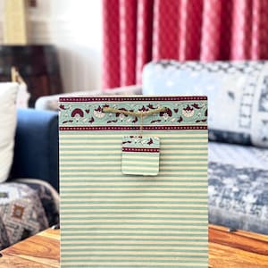 May include: A rectangular gift bag with a striped pattern in light blue and cream. The top border features a floral design in burgundy and cream. A matching tag is attached with twine. The bag is sitting on a wooden surface.
