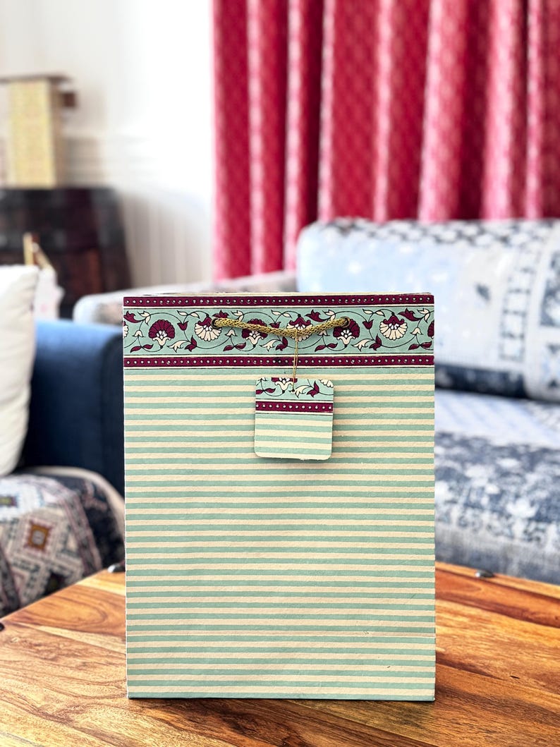 May include: A rectangular gift bag with a striped pattern in light blue and cream. The top border features a floral design in burgundy and cream. A matching tag is attached with twine. The bag is sitting on a wooden surface.