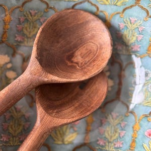 Op de afbeelding: Twee houten lepels, diep bruin van kleur, liggen op elkaar. Ze rusten op een turquoise oppervlak versierd met een bloemenpatroon in geel, roze en wit.