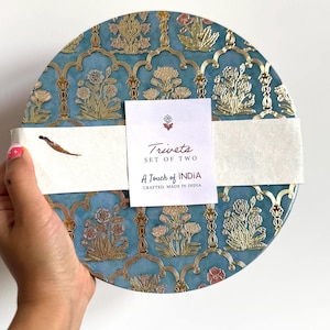 May include: A pair of round trivets featuring a blue and gold floral pattern. The trivets are wrapped with a white band and a tag that reads "Trivets SET OF TWO A Touch of INDIA CRAFTED. MADE IN INDIA."